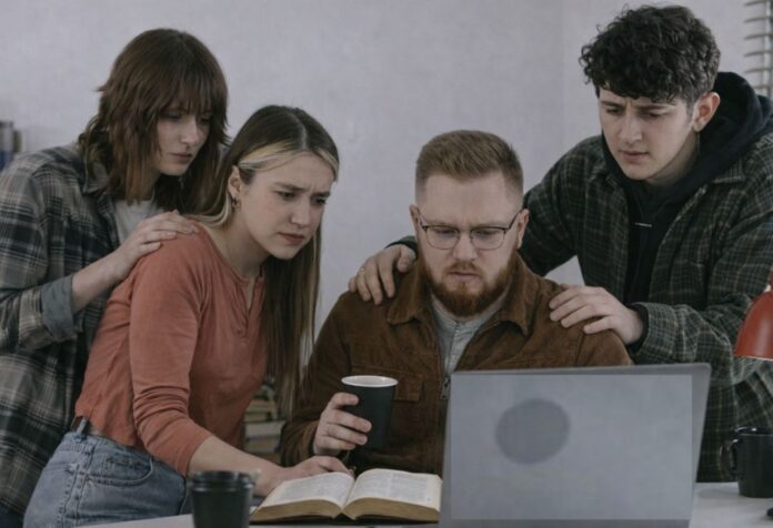 Cuatro jóvenes observan con preocupación las noticias en una computadora portátil, mientras se acompañan con gestos de cercanía; una Biblia abierta permanece sobre el escritorio.