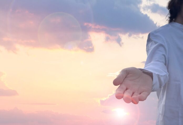 Figura de Jesús con túnica blanca extendiendo la mano hacia el espectador, con un cielo iluminado al amanecer de fondo.