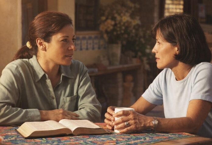 Dos mujeres conversan con atención en un ambiente cálido de hogar, con una Biblia abierta sobre la mesa, reflejando diálogo respetuoso y profundidad espiritual.