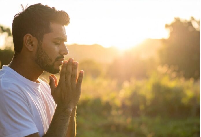 Hombre con los ojos cerrados y las manos juntas en oración al amanecer, iluminado por la luz del sol.