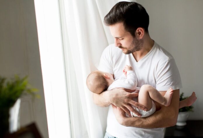Un padre sostiene con ternura a un bebé en brazos junto a una ventana iluminada. La escena transmite cercanía, cuidado y el vínculo afectivo entre padre e hijo.